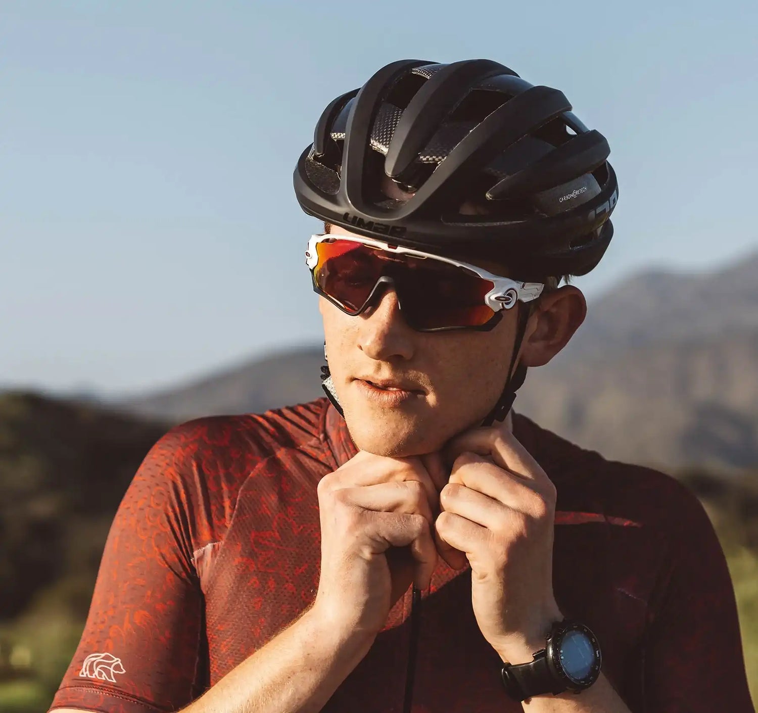Cyclist adjusts black Limar helmet, wearing sunglasses and jersey with mountain backdrop